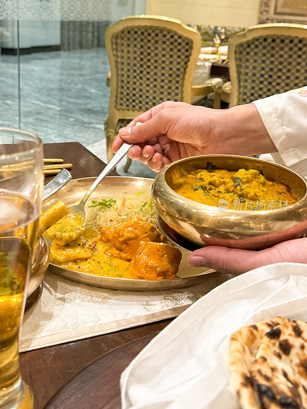 特写照片中，无法辨认的服务员用勺子从金属碗里端上Dal makhani(黑扁豆和红芸豆咖喱)，餐厅背景，重点放在前景上